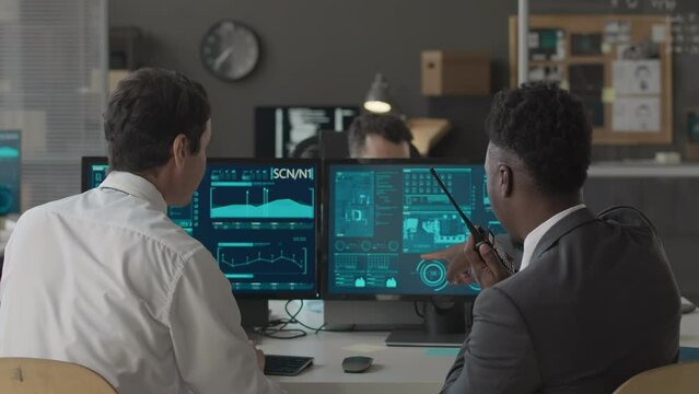 Dolly-out back-view shot of ethnically diverse male colleagues co-working in cybersecurity agency, looking at secret network data program and transmitting info through walkie talkie