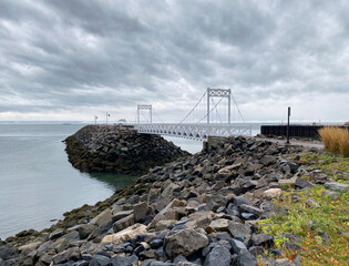 Pier on the coast. Structure of a pedestrian bridge on a quay. Walkway at the river's edge on a cloudy day. White gate on a rocky bank. Boat and buoy in the distance.