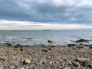 Beach and rocks on a cloudy day. Coastal beach at low tide. Stones on the coast. Cargo boat in the distance on the river.