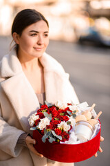 Young smiling woman holding a gift box with an arrangement of roses along with an aromatic candle and macaroons
