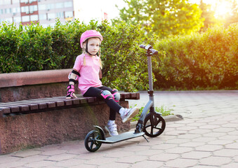 A beautiful seven-year-old girl sits on a bench in pink protective equipment next to a scooter. The concept of cycling and scootering without injury, sunset