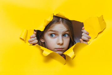 Funny red-haired child girl peeping through hole on yellow paper. The concept of surprise, fear, fright, joyful mood from what he saw. Discounts, sales, surprise. Copy space.