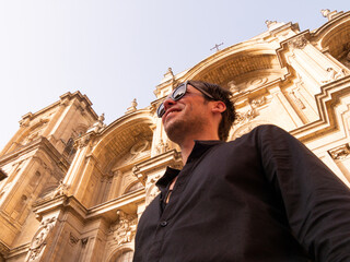 Caucasian man tourist with open black shirt and sunglasses smiles in front of the Granada cathedral looking up at the horizon to the side - 768850988