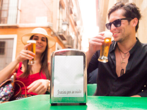unfocused couple laughing drinking beer behind a napkin holder focused on the one that says thank you for your visit on a bar terrace