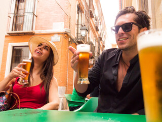 straight hispanic caucasian couple with sunglasses and dental braces hat and black shirt red tshirt laughing drinking beer on a terrace of a spanish bar pointing up - 768851302