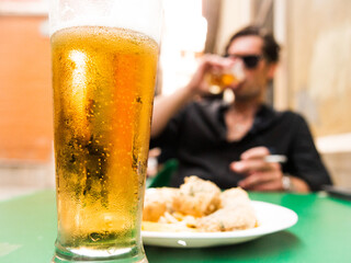 glass of beer on Spanish bar terrace with defocused caucasian man drinking and with cigar and sunglasses in background and fried fish appetizer - 768852148
