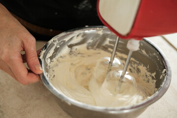 A close-up of the hands of a young woman pastry chef who is whipping butter cream for a cake using an electric mixer in a deep iron bowl. The process of preparation in production in a confectionery.