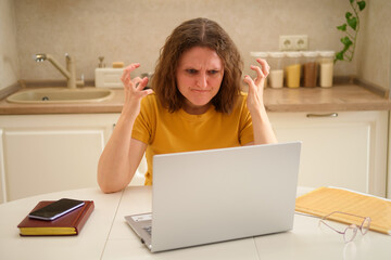 Angry woman with a laptop is sitting at a table in a home kitchen. An adult female businesswoman...