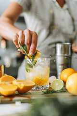 An action shot of a refreshing citrus cocktail being garnished with rosemary, surrounded by fresh lemons and limes.
