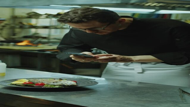 Chef taking photos with mobile phone of fish served with vegetables on plate in kitchen, preparing social media content for restaurant. Vertical clip