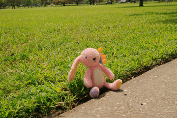 Pink stuffed rabbit sitting in green grass beside the sidewalk. Purple and yellow feet with yellow...