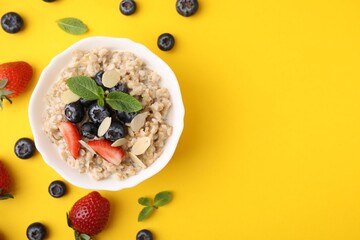 Tasty oatmeal with strawberries, blueberries and almond petals in bowl surrounded by fresh berries on yellow background, flat lay. Space for text