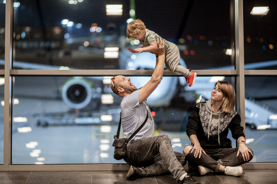 Riga, Latvia - October 19, 2019 - A man lifts a laughing toddler into the air while sitting beside a woman at an airport, with a plane visible through the window behind them.