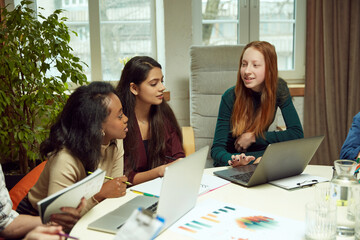 Obraz premium Young girls, friends, students sitting at table with papers and laptops, studying, finishing tasks together. Concept of education, youth, knowledge, homework, exams, projects.
