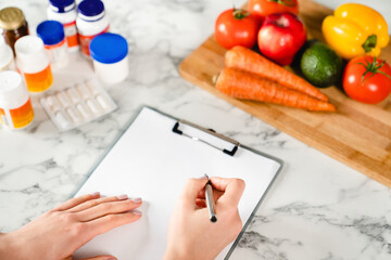 Close up shot of woman`s hands writing on clipboard close to the food supplements and fresh...