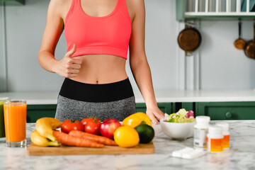 Cropped shot of slim sporty woman showing thumb up at kitchen table with organic vegetables and...