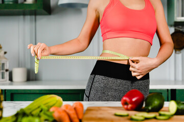 Young sport girl measuring her waist with measuring tape in modern kitchen with vegetables on...