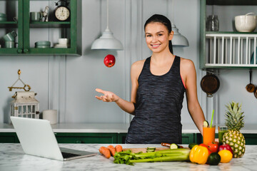 Fit young woman lifting an apple up in the kitchen using laptop. Healthy eating concept. Veganism...