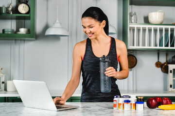 Healthy fit young woman using laptop for additional info about food supplements in the kitchen....