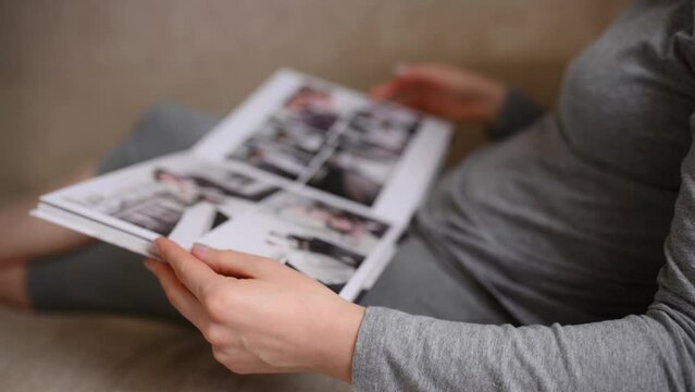 Close-up of a woman leafing through large wedding photo book at home, lying on the sofa in living room. Concept of photo printing, positive memories