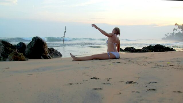 Woman in bikini performs side bow sitting in leg-split on sandy beach by ocean. Lady with braids enjoys yoga session at sunset