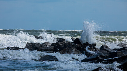 Waves crash on dark rocks with a uniform blue sky above.  White Sea foam and ocean spray and small waves in the foreground.  Waves crash during high tide in Florida on the Atlantic Ocean coastline.