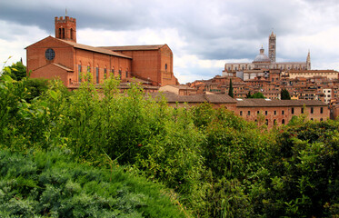 Naklejka premium Panoramic view of the historic part of the city of Siena with the Duomo di Siena and green trees and bushes in the foreground against a stormy sky with clouds in the Tuscany region of Italy