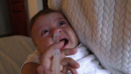 Removing baby mucus by decongesting nose with saline syringe. Mother holding crying baby upset by...