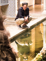 Thirty-year-old man looks smiling at himself in the reflection of a fountain while crouching - 768920556