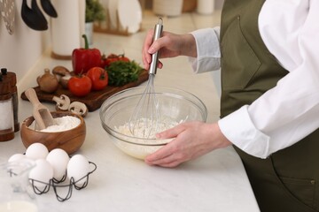 Woman making dough with whisk in bowl at light table, closeup