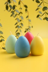 Fototapeta na wymiar Colorful egg shaped candles and leaves on yellow background. Easter decor