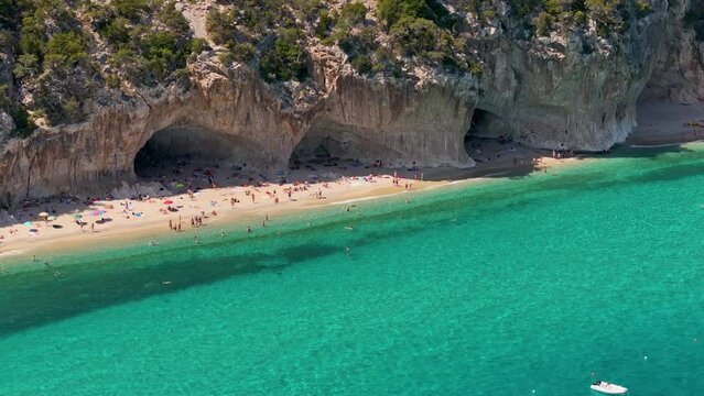 Aerial shot - Huge rocks on the coastline giving shade on a beautiful sandy beach crowded with tourist on summer vacations. Cala Luna, Sardinia, Italy.