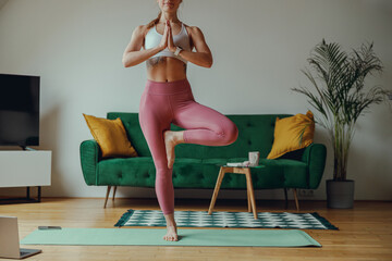 A woman in sportswear is balancing on one leg on a yoga mat in a living room