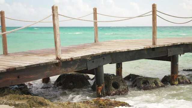 Waves crashing by the concrete reef balls under an empty wooden pier on a tropical hotel beach with turquoise water of Caribbean sea. Cloudy stormy day relax background