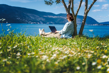 Young woman sitting on a green summer lawn on the seashore using a laptop - 768945999