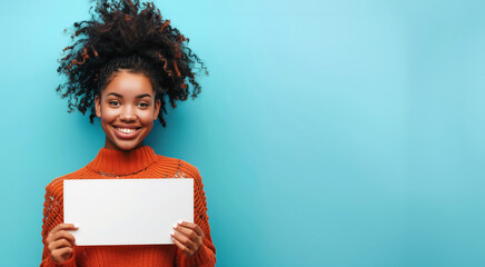 A young smiling Black woman in an orange sweater holds up a white blank paper sheet on a blue background, banner for advertising copy space, blank sign for your advertising text messages - obrazy, fototapety, plakaty