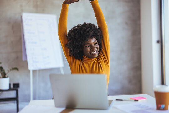Happy carefree African American woman stretching hands at workplace, leaning back in comfortable chair, student or freelancer relaxing enjoying break after work done, sitting at desk with laptop