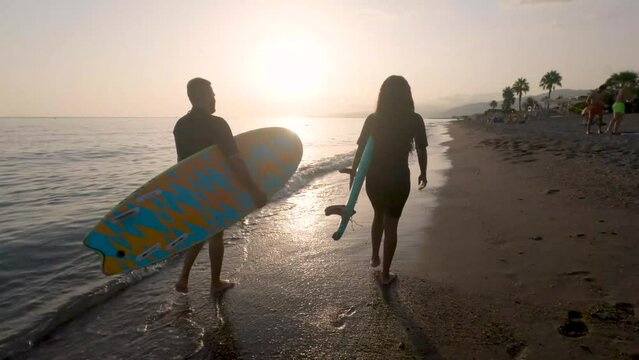 A young multi-ethnic couple stroll along the shore of the beach at sunset with south boards. African man and woman talk happily as they walk.Water sport concept.