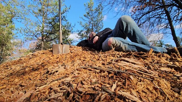 Man in glasses wearing jacket and jeans taking break on top of mulch laying on pile next to impaled shovel or spade after working in his garden.