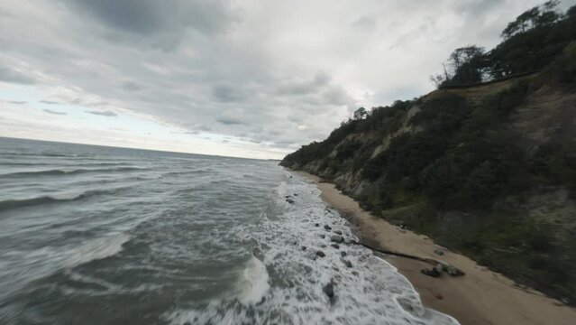 FPV drone flight above picturesque stormy foamy ocean waves washing rocky coast with cliffs under gloomy sky in overcast weather