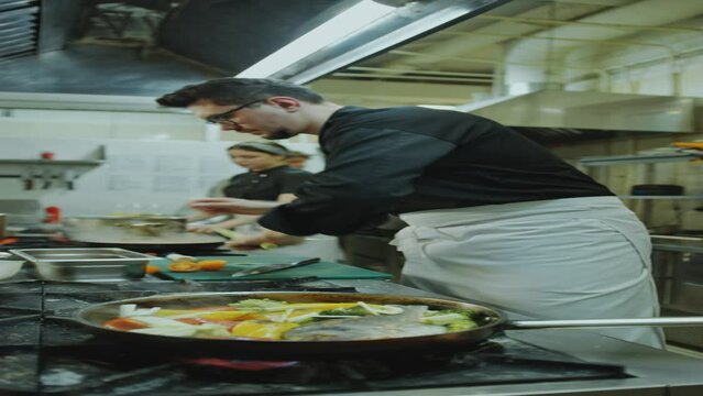 Chef adding leek and cherry tomatoes to fish with vegetables in cooking pan on stove while preparing food at work in restaurant kitchen. Vertical clip