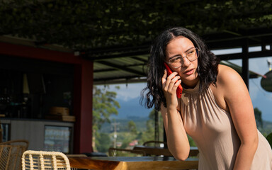 mujer joven latina de cabello corto y usando lentes mientras habla por teléfono y mira a un...