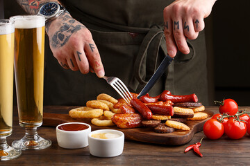 Set of assorted beer snacks - grilled sausages, roasted chicken wings, croutons, deep fried onion rings and squid rings on wooden table with glass of beer and sauces. Male hands cuts sausage