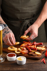 Set of assorted beer snacks - grilled sausages, roasted chicken wings, croutons, deep fried onion rings and squid rings on wooden table with glass of beer and sauces. Chef serving a dish 