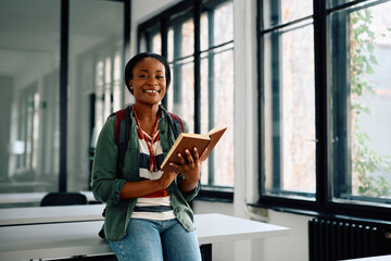 Happy black university student reading  book in classroom and looking at camera.