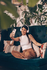 A young dark-haired woman in casual clothes sitting on the couch,wearing headphones,using a smartphone.Relaxation,meditation,listening music,taking a selfie.