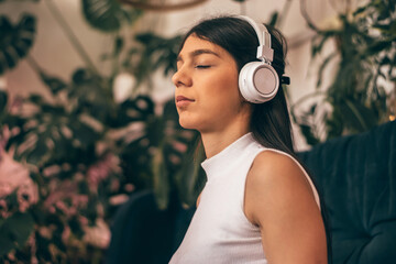 A young dark-haired woman in casual clothes wearing headphones,sitting on the couch with closed eyes.Relaxation,meditation,listening music.