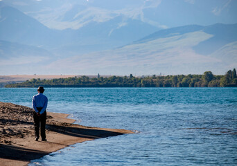 Naklejka premium An old man walking by the shore of the famous Issyk-Kul lake surrounded by the Ala-Too mountains. Kyrgyzstan, July 2023