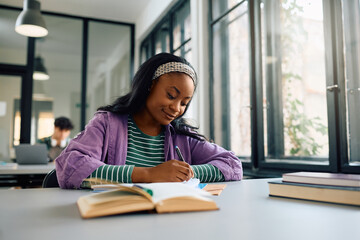 Black female student writing notes while learning in  library.