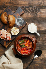 Traditional Russian and Ukrainian beet soup borsch with beef and mushrooms in clay pot served with rye bun, bacon, garlic, sour cream and glass of vodka on wooden table. Overhead view